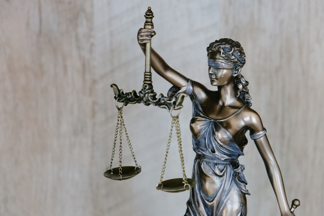 A bronze statue of Lady Justice holding a balance scale in her left hand and a sword in her right hand. She is blindfolded, symbolizing impartiality. The background is blurred and neutral.